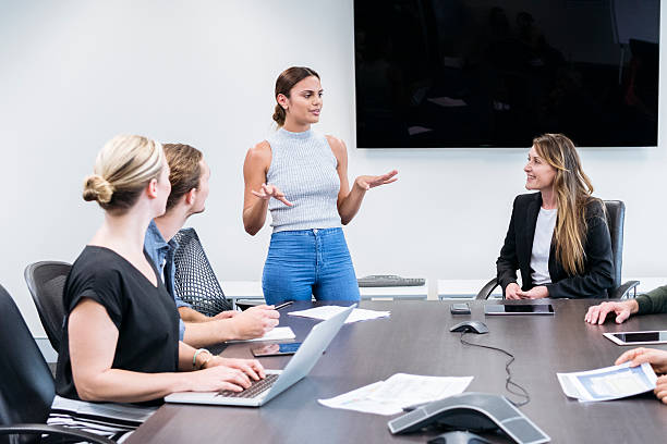 Casual businesswoman standing by table in meeting room, colleagues listening, woman using laptop